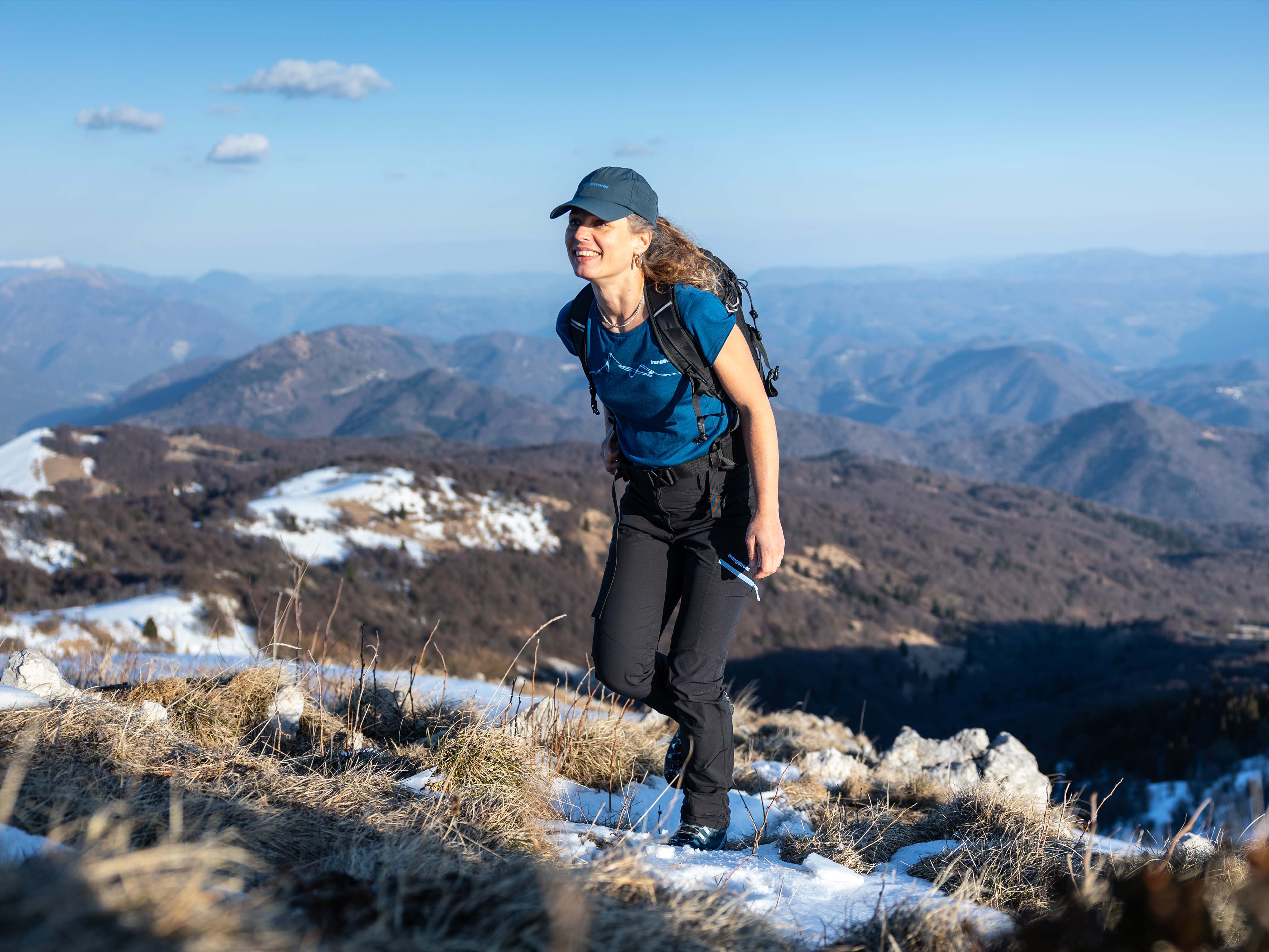 Senderista con gorra caminando por cresta nevada Mujer con camiseta de lana merino Trangoworld azul haciendo senderismo en montaña con nieve
