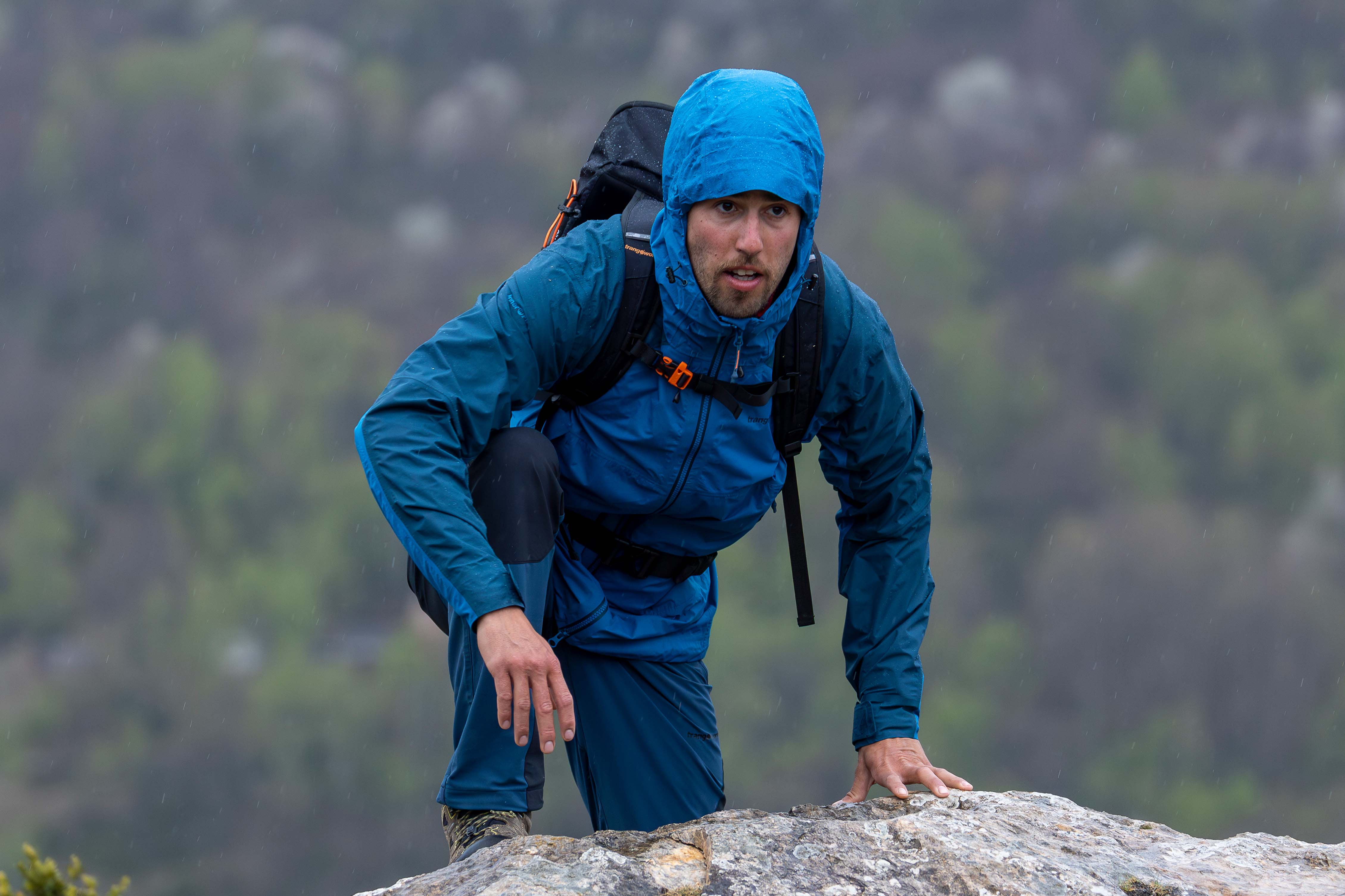 Chaqueta impermeable técnica para montaña. Cortavientos. Trangoworld Hombre con chaqueta impermeable azul Trangoworld con mochila bajo la lluvia